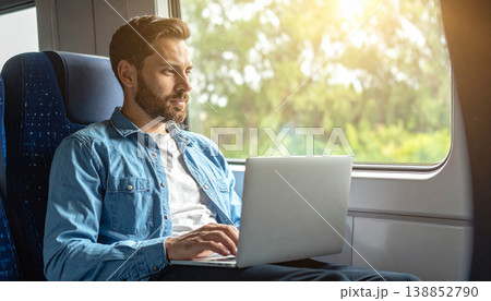 Young man working on laptop during train travel at sunset. Professional digital nomad lifestyle with golden sun flare, remote working, and commuter journey in modern railway carriage interior 138852790