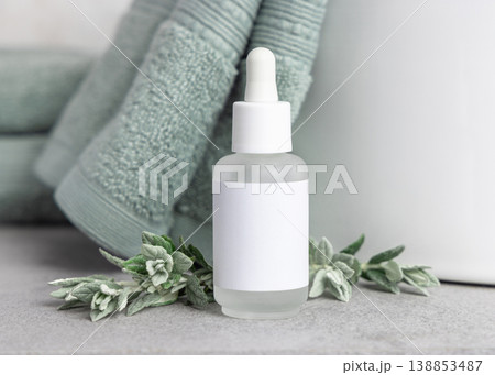 Dropper bottle with blank label mockup near leaves and sage green bath towel. Cosmetic packaging 138853487