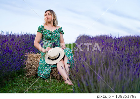 Woman in floral dress sits amidst a lavender field, holding a straw hat. Woman in floral dress sits amidst a lavender field, holding a straw hat. 138853768