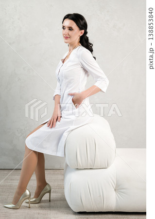 Elegant woman in white lab coat sits gracefully on a modern white sofa, showcasing professionalism and poise. Elegant woman in white lab coat sits gracefully on a modern white sofa, showcasing professionalism and poise. 138853840