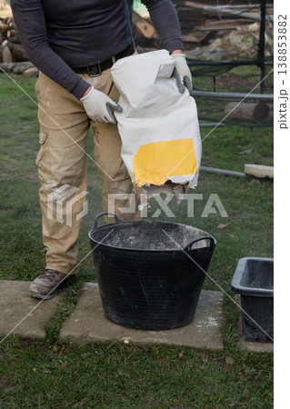 Person pours material self-leveling floor from a bag into a black bucket while standing on a path in a yard during daylight 138853882