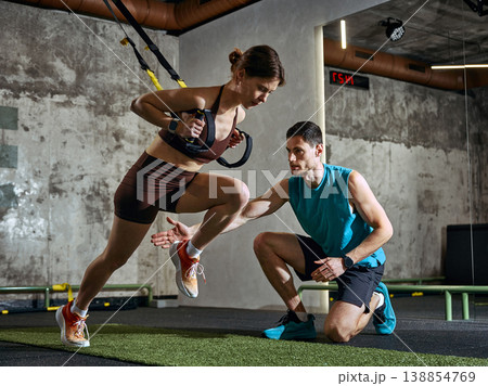 Woman training with suspension straps while man guiding movement in gym Woman training with suspension straps while man guiding movement in gym 138854769