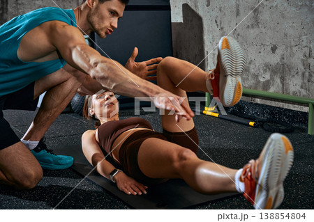 Woman performing leg raise exercise on mat with male coach guiding technique in gym. Woman performing leg raise exercise on mat with male coach guiding technique in gym. 138854804
