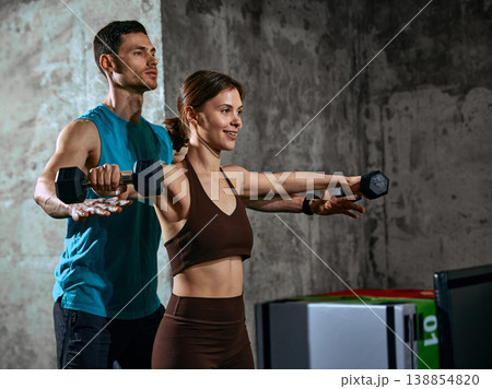Woman doing dumbbell lateral raise exercise with male instructor correcting form in gym. Woman doing dumbbell lateral raise exercise with male instructor correcting form in gym. 138854820