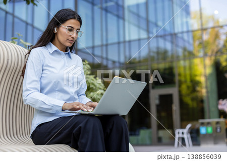 Young businesswoman typing on a laptop computer while sitting on a modern bench outside a glass office building, embracing flexible work and global connectivity Young businesswoman typing on a laptop computer while sitting on a modern bench outside a glass office building, embracing flexible work and global connectivity 138856039