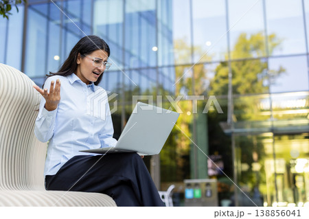 Young business woman experiencing stress and frustration while working on a laptop computer outside a modern office building, encountering a technical error or failure 138856041