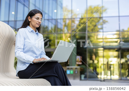 Young indian businesswoman sitting on a contemporary bench outside a corporate glass building, typing on her laptop, focusing on work and productivity in a professional urban setting 138856042