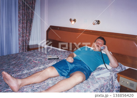 Man lying on hotel bed talking on landline phone 1990s 138856420