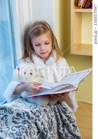 A cute baby in the room sits at the window in the winter and reads a book A cute baby in the room sits at the window in the winter and reads a book 138857097