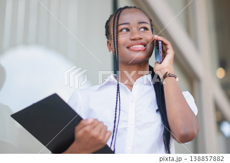 Young Black business woman holding a tablet and making a smartphone call outside, smiling confidently. Remote work, mobile communication, multitasking and modern professional lifestyle concept. 138857882