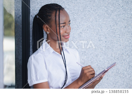 Young Black business woman leaning against a wall while writing on a tablet outdoors. Mobile workflow, digital planning, field notes, remote work and modern productivity concept. Young Black business woman leaning against a wall while writing on a tablet outdoors. Mobile workflow, digital planning, field notes, remote work and modern productivity concept. 138858155
