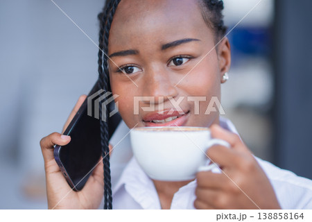 Close-up of young Black woman talking on smartphone while holding coffee in a cafe. Friendly mobile communication, customer support, remote business contact and modern lifestyle concept. Close-up of young Black woman talking on smartphone while holding coffee in a cafe. Friendly mobile communication, customer support, remote business contact and modern lifestyle concept. 138858164