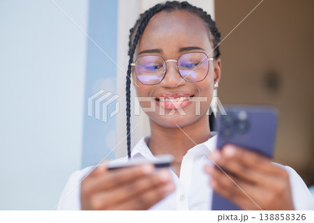 Smiling young Black woman in glasses holding credit card and smartphone for online payment. Mobile banking, digital shopping, contactless finance, ecommerce transaction and fintech lifestyle concept. 138858326