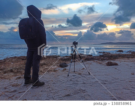 Male Photographer Standing with Hands in Pockets by DSLR on Tripod at Sunset on Beach Male Photographer Standing with Hands in Pockets by DSLR on Tripod at Sunset on Beach 138858961