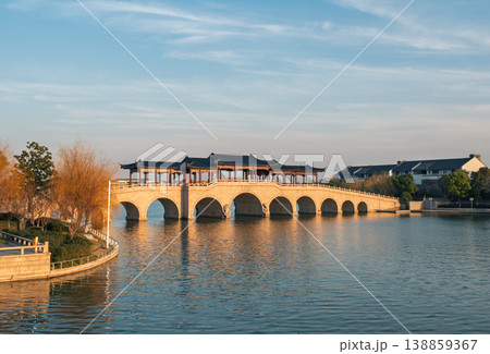 Covered stone arch bridge with multiple spans extends across a placid lake during sunset. Covered stone arch bridge with multiple spans extends across a placid lake during sunset. 138859367