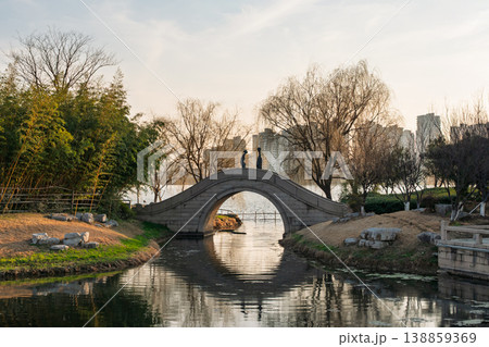 Stone arch bridge with two stylized figures arches over a peaceful canal. 138859369