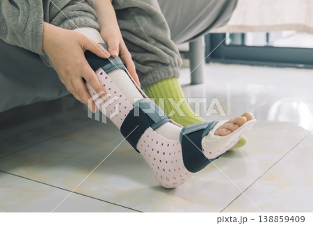 Close up of a person adjusting a medical ankle splint on their injured foot indoors. Close up of a person adjusting a medical ankle splint on their injured foot indoors. 138859409