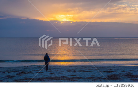 Silhouette of Man Walking Toward the Shore at Sunset with Calm Sea and Cloudy Sky 138859916