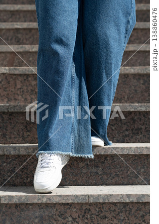 Low angle view of person wearing wide leg blue jeans and white sneakers walking down granite stairs. 138860476