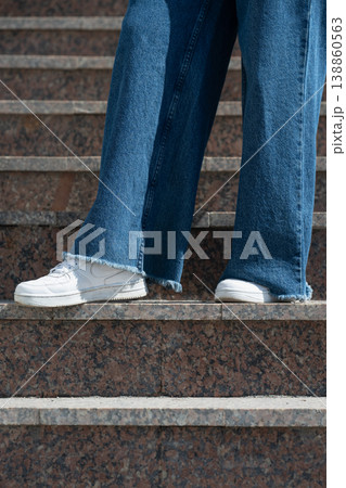 Side view of person wearing wide leg blue jeans with raw hem and white sneakers on granite stairs. 138860563