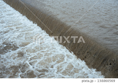 Turbulent muddy river water flowing over a small dam with white foam and splashes. Turbulent muddy river water flowing over a small dam with white foam and splashes. 138860569