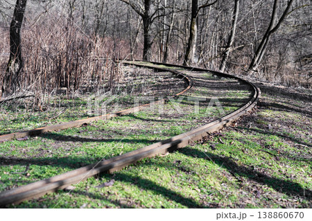 Abandoned railway tracks curving through a quiet forest in early spring. Old railroad rails covered with moss and grass, surrounded by bare trees and dry bushes. Abandoned railway tracks curving through a quiet forest in early spring. Old railroad rails covered with moss and grass, surrounded by bare trees and dry bushes. 138860670