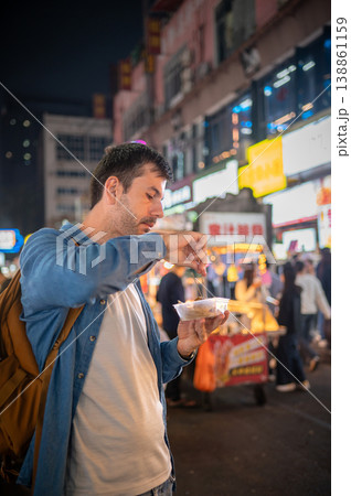 Man Enjoying Street Food at Vibrant Night Market in China, Surrounded by Colorful Neon Lights and Bustling Atmosphere 138861159