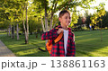 Joyful teenager walking in sunny park with orange backpack, smiling. Youth lifestyle, casual outdoor activity, summer, leisure, calm and happy everyday moment. Teenage schoolboy at sunset or sunrise. 138861163
