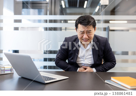 Asian businessman sitting at office desk feeling severe stomach pain and discomfort, holding his stomach while grimacing in distress from indigestion or a health issue 138861481