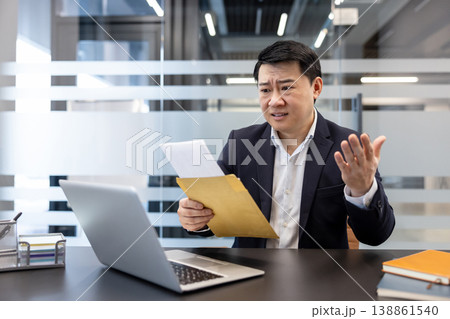 Asian businessman in suit at desk, reacting with worry and hand gestures while opening an envelope and reading a troubling document beside his laptop in a modern office Asian businessman in suit at desk, reacting with worry and hand gestures while opening an envelope and reading a troubling document beside his laptop in a modern office 138861540