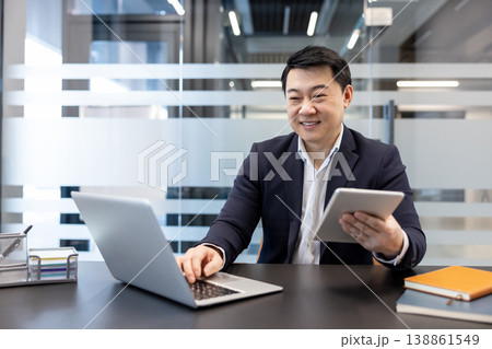 Asian businessman performing digital work, smiling while multitasking with a laptop and digital tablet, completing tasks and managing information efficiently in a modern office environment 138861549