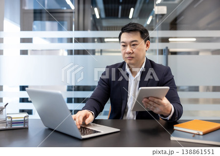 Asian businessman working in a corporate office. Simultaneously typing on a laptop and holding a digital tablet. Demonstrating multitasking and technological integration in a professional environment 138861551