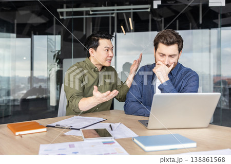 Two diverse male business colleagues engaging in a heated argument at an office desk, one man expressing anger while the other appears frustrated and stressed Two diverse male business colleagues engaging in a heated argument at an office desk, one man expressing anger while the other appears frustrated and stressed 138861568