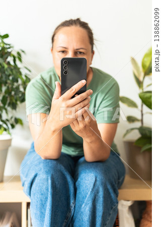 An adult woman focusing on phone screen, holding hand on chin, thinking about information or making a decision. Chatting, surfing social media, watching video content 138862099