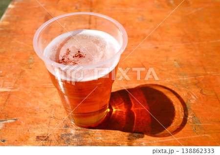 Closeup of plastic cup of cold beer with foam on orange wooden table at outdoor summer event 138862333