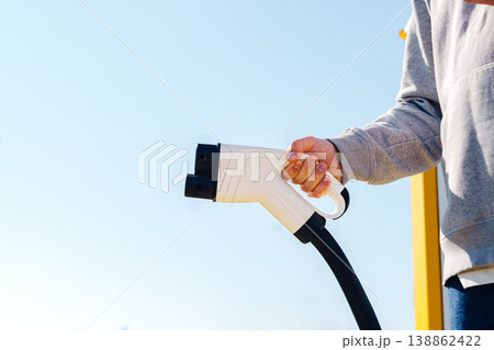 Person holds electric car charger in hands at outdoor charging station in sunlight Person holds electric car charger in hands at outdoor charging station in sunlight 138862422
