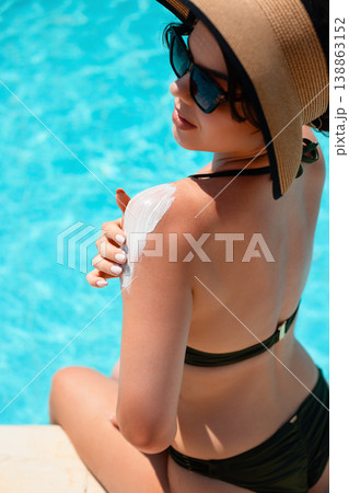 Woman in black bikini and straw hat sitting by pool from back angle applying sunscreen on shoulder enjoying bright sunlight and relaxed summer vacation mood Woman in black bikini and straw hat sitting by pool from back angle applying sunscreen on shoulder enjoying bright sunlight and relaxed summer vacation mood 138863152