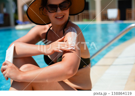 Woman Smiling While Applying Sunscreen On Shoulder Sitting By Poolside In Summer Sunlight. Skincare And Sun Protection Concept Woman Smiling While Applying Sunscreen On Shoulder Sitting By Poolside In Summer Sunlight. Skincare And Sun Protection Concept 138864119