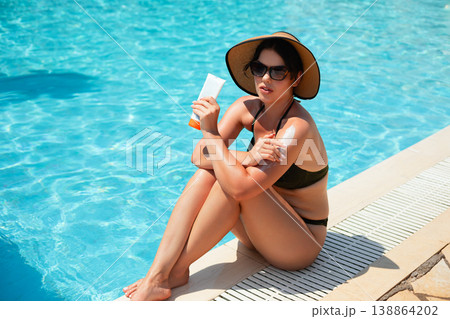 Woman sitting by pool holding sunscreen and applying on shoulder wearing sunglasses and hat enjoying bright sunny summer vacation lifestyle Woman sitting by pool holding sunscreen and applying on shoulder wearing sunglasses and hat enjoying bright sunny summer vacation lifestyle 138864202