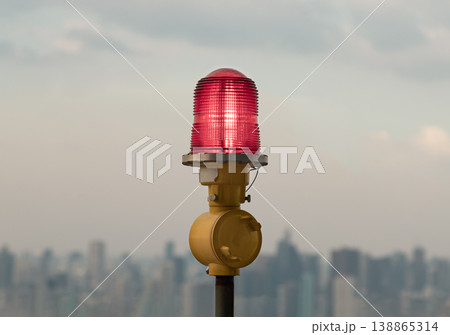 Against a hazy, metropolitan horizon, a singular red obstruction light or aircraft warning light stands as a silent sentinel. The beacon, encased in a crimson, ribbed glass housing, glows with a soft Against a hazy, metropolitan horizon, a singular red obstruction light or aircraft warning light stands as a silent sentinel. The beacon, encased in a crimson, ribbed glass housing, glows with a soft 138865314