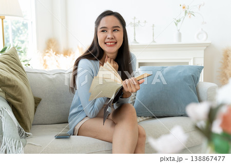 Joyful Asia woman Relaxing on Sofa with book diary. A young woman finds peace and tranquility as she relaxes on a comfortable sofa, immersed in the captivating world of literature. Lifestyle Concept Joyful Asia woman Relaxing on Sofa with book diary. A young woman finds peace and tranquility as she relaxes on a comfortable sofa, immersed in the captivating world of literature. Lifestyle Concept 138867477