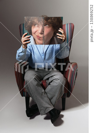 Young man sitting in armchair holding notebook covering face minimal studio portrait. Young man sitting in armchair holding notebook covering face minimal studio portrait. 138869252