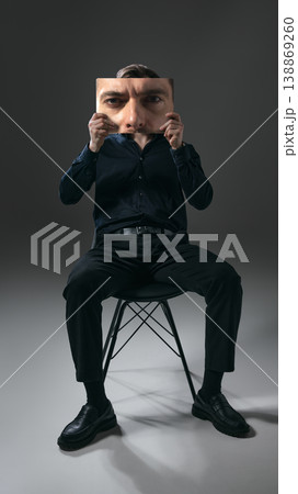 Man in black outfit holding closeup face print while sitting on chair minimal studio portrait. 138869260