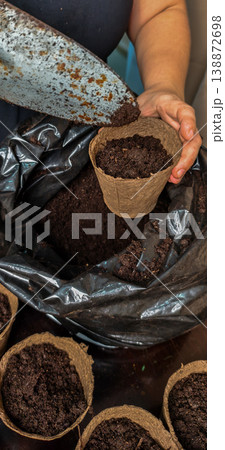 Woman's hands using a gardening trowel to fill an eco-friendly biodegradable pot with dark soil, preparing it for planting vegetable or flower seedlings, promoting sustainable home gardening. Woman's hands using a gardening trowel to fill an eco-friendly biodegradable pot with dark soil, preparing it for planting vegetable or flower seedlings, promoting sustainable home gardening. 138872698