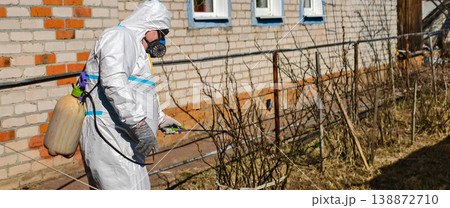 Worker wearing a protective chemical suit and respirator mask, actively spraying dormant garden bushes with a manual sprayer, preparing them for the growing season. Worker wearing a protective chemical suit and respirator mask, actively spraying dormant garden bushes with a manual sprayer, preparing them for the growing season. 138872710
