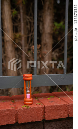 Vintage orange hourglass on red brick ledge by forest fence Vintage orange hourglass on red brick ledge by forest fence 138873582