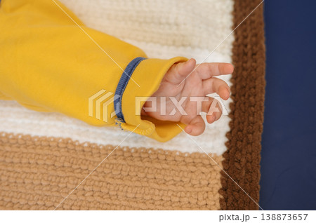 Baby hand resting on striped crocheted blanket beside yellow sleeve on blue Baby hand resting on striped crocheted blanket beside yellow sleeve on blue 138873657