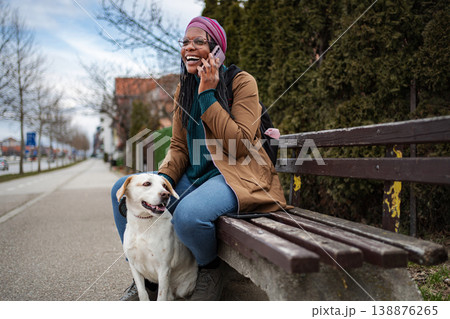 Happy woman talking on phone with dog at park bench 138876265