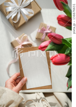 Vertical shot of woman's hands holding a blank white card over table with red tulips and craft gifts. Greeting card mockup Vertical shot of woman's hands holding a blank white card over table with red tulips and craft gifts. Greeting card mockup 138876631