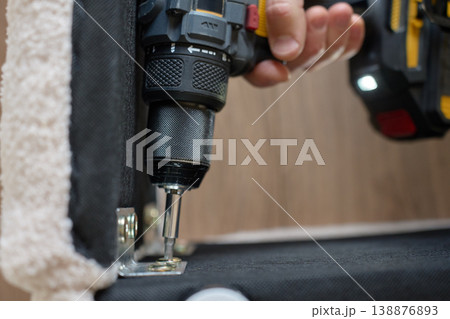 Cordless screwdriver fastening screw into metal bracket on upholstered furniture frame 138876893